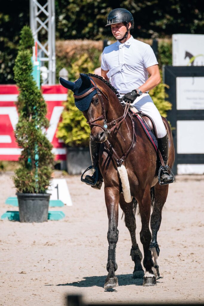 Reiter in weißem Outfit auf braunem Pferd mit blauer Kappe, konzentrierter Ausdruck, sonniger Tag.