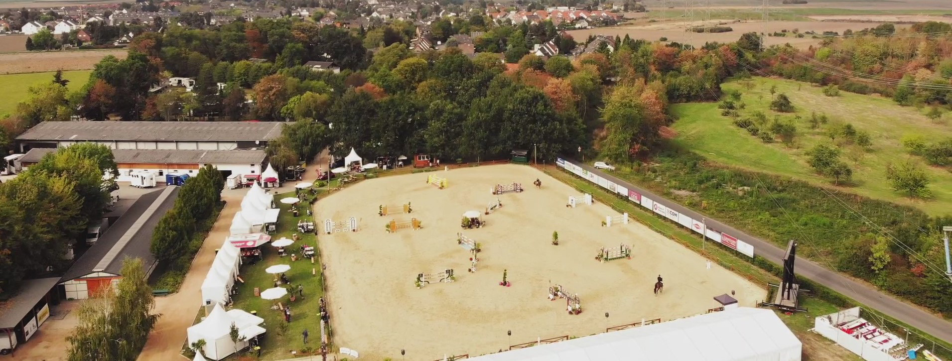 Luftaufnahme eines Reitturniers mit Springhindernissen, Zelten und umliegenden Bäumen.