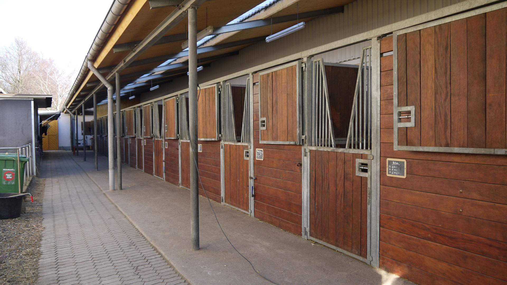 Eine Reihe von Pferdeboxen aus Holz in einem Stall mit überdachtem Gang.