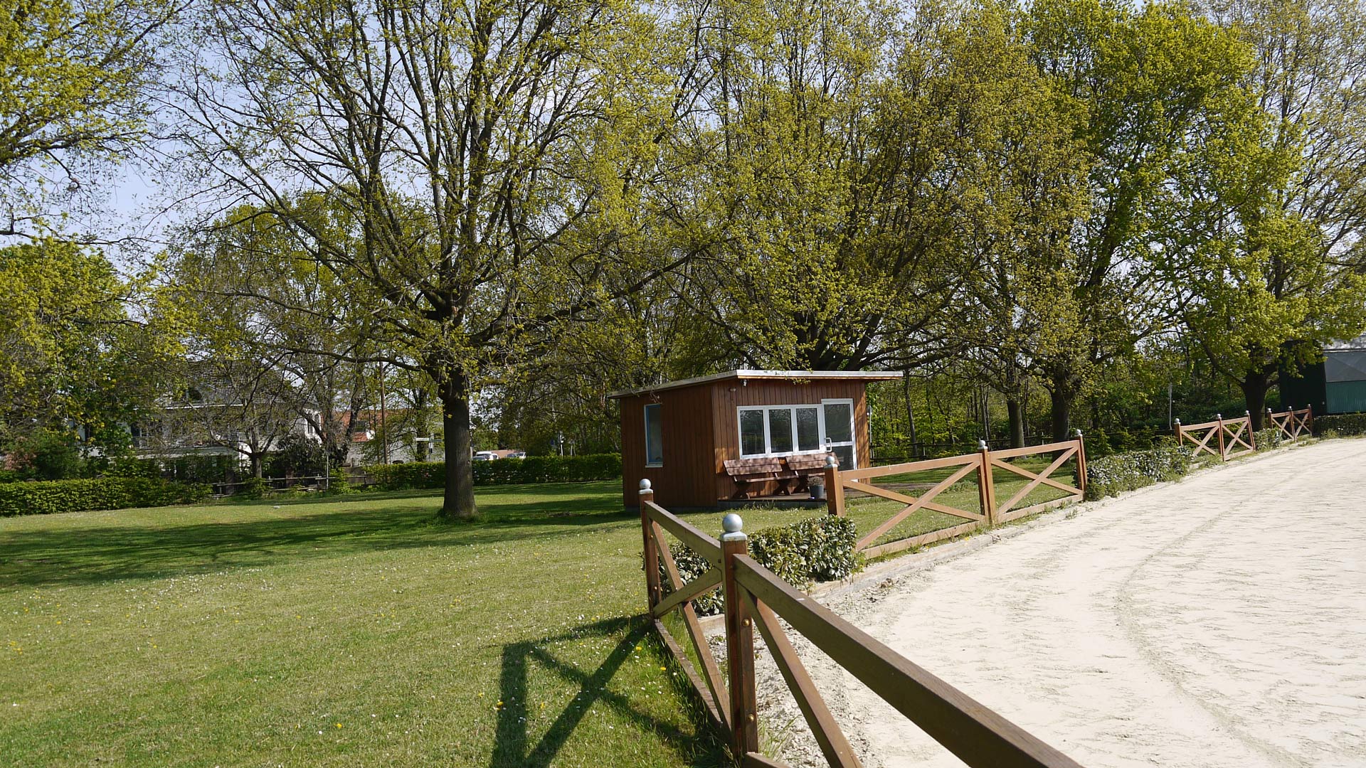 Eine kleine, braune Holzhütte in einem grünen Park mit Bäumen und einem Holzzaun im Vordergrund.