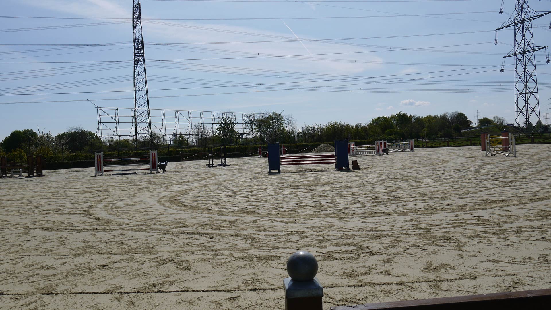 Weitläufiger, leerer Reitplatz mit Sprunghindernissen und Strommasten im Hintergrund.