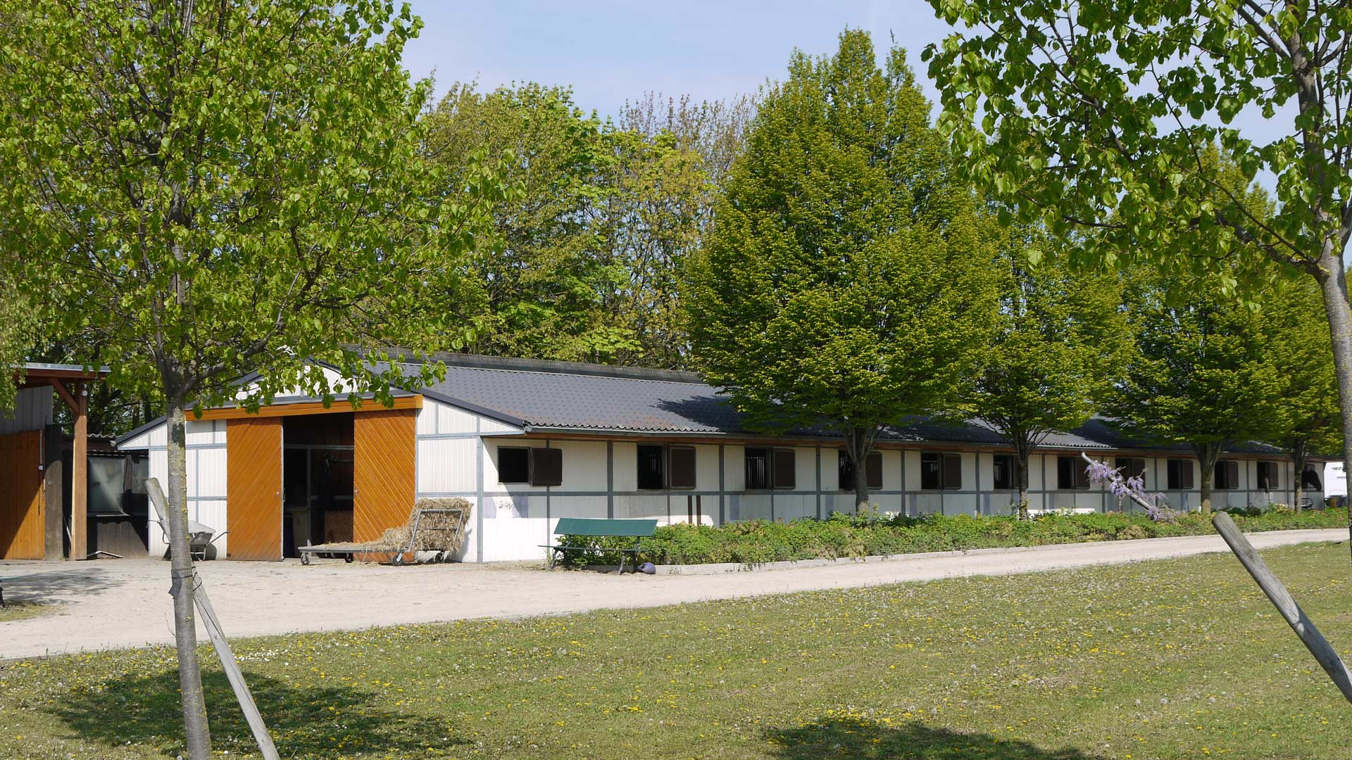 Ein langes, weißes Gebäude mit Holztüren, umgeben von grünen Bäumen unter blauem Himmel.