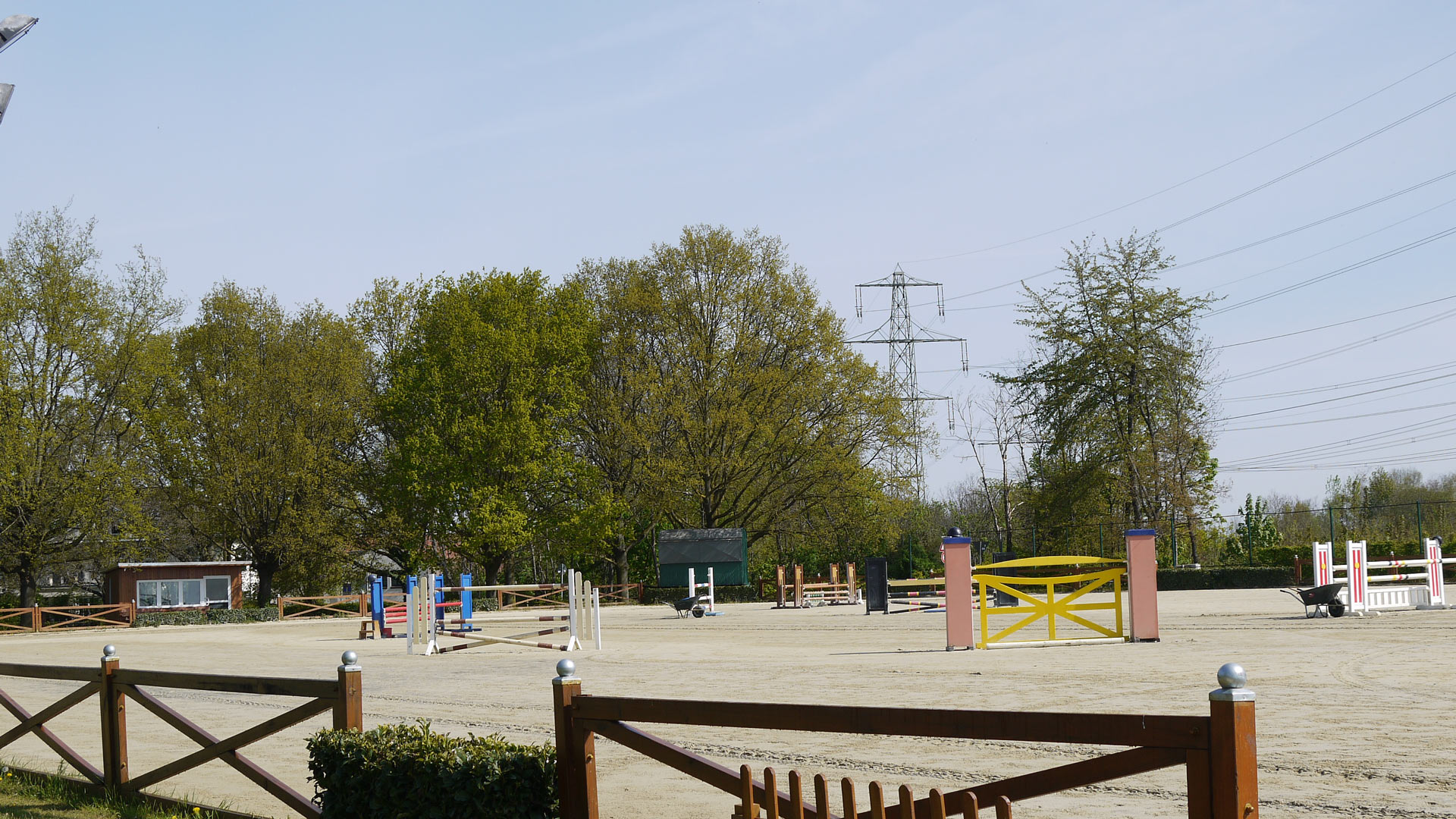 Ein leerer Reitplatz mit bunten Hindernissen, umgeben von Bäumen und einem Holzzaun.