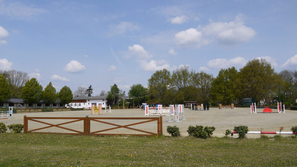 Eine leere Reitplatzszene mit Sprunghindernissen, grüne Bäume und blauer Himmel im Hintergrund.