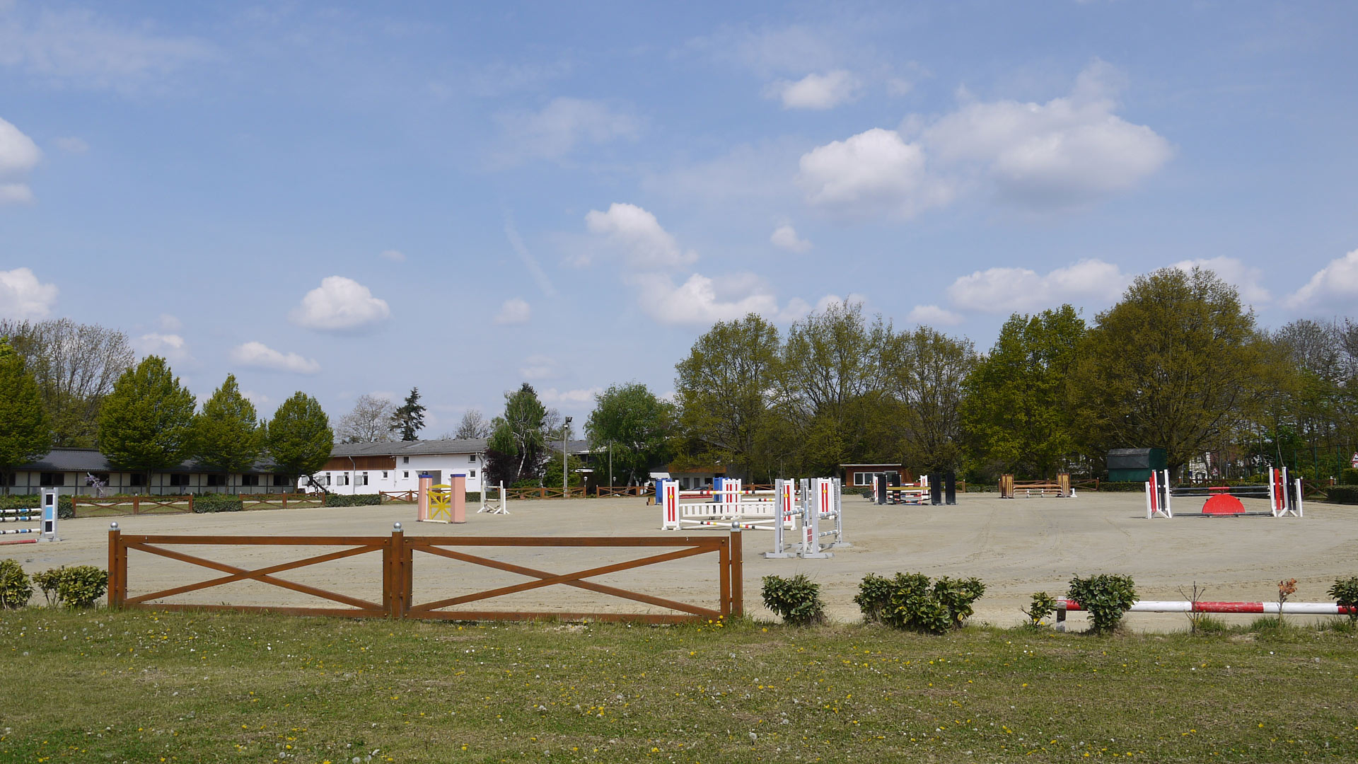 Reitplatz mit Hindernissen, umgeben von Bäumen und Gebäuden unter blauem Himmel mit Wolken.