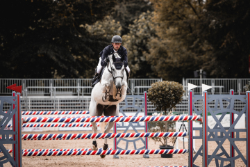Person in Reitkleidung springt mit weißem Pferd über rot-blau gestreiftes Hindernis im Freien.
