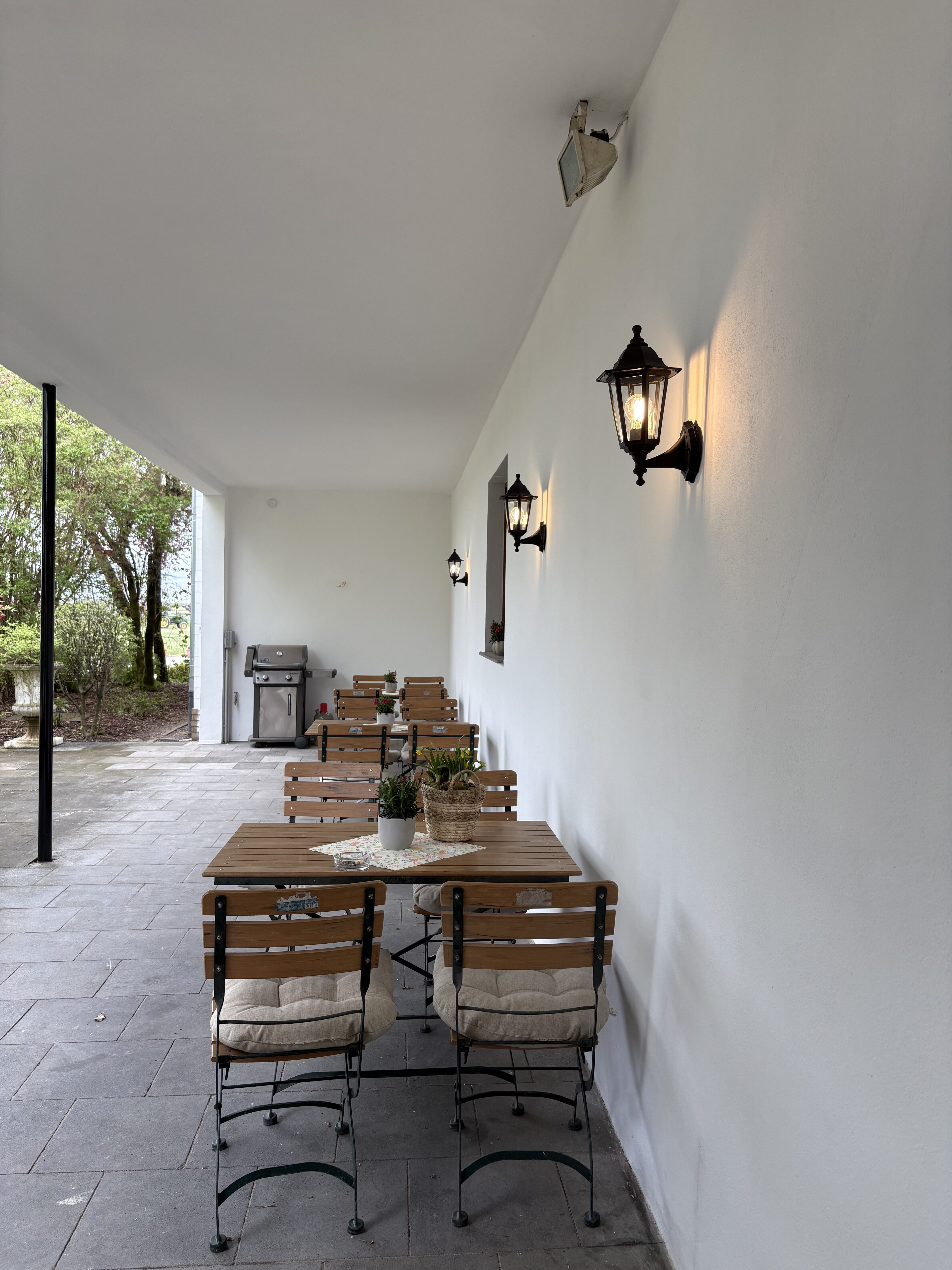 Ueberdachte Terrasse mit Holztischen Stuehlen und Laternen an weisser Wand